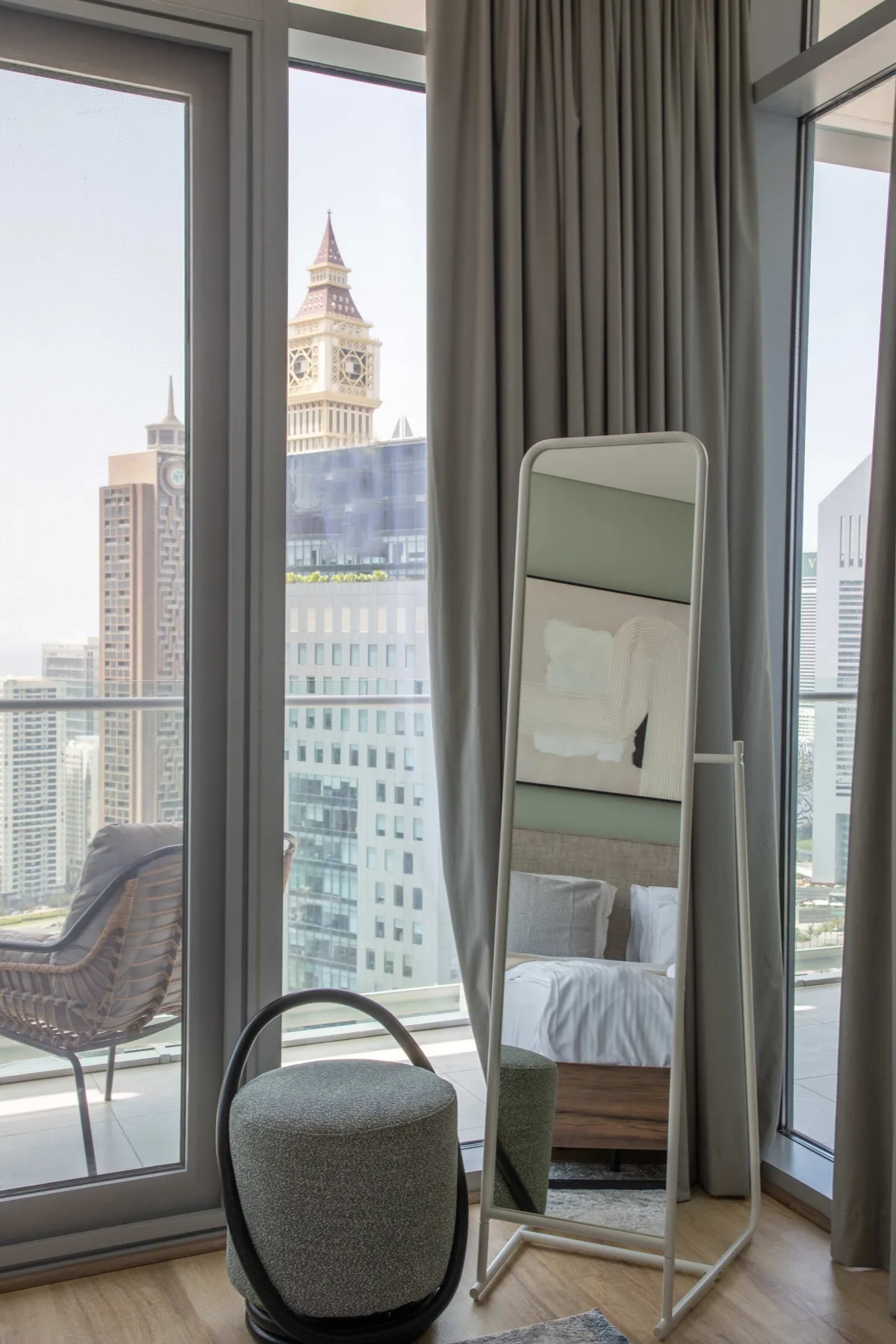 A bedroom with a floor mirror beside floor-to-ceiling windows overlooking city buildings.