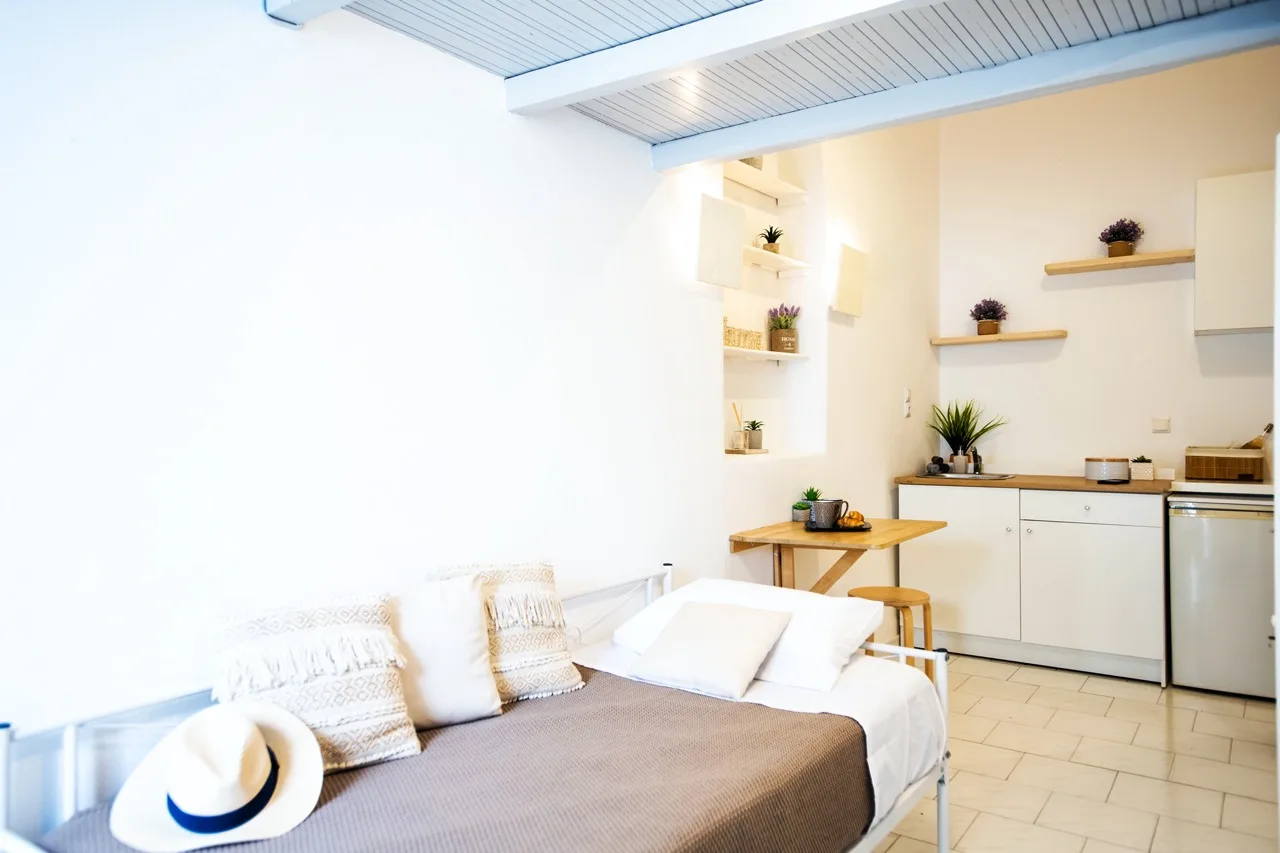 A bright studio room with a bed, small dining table, and kitchenette.