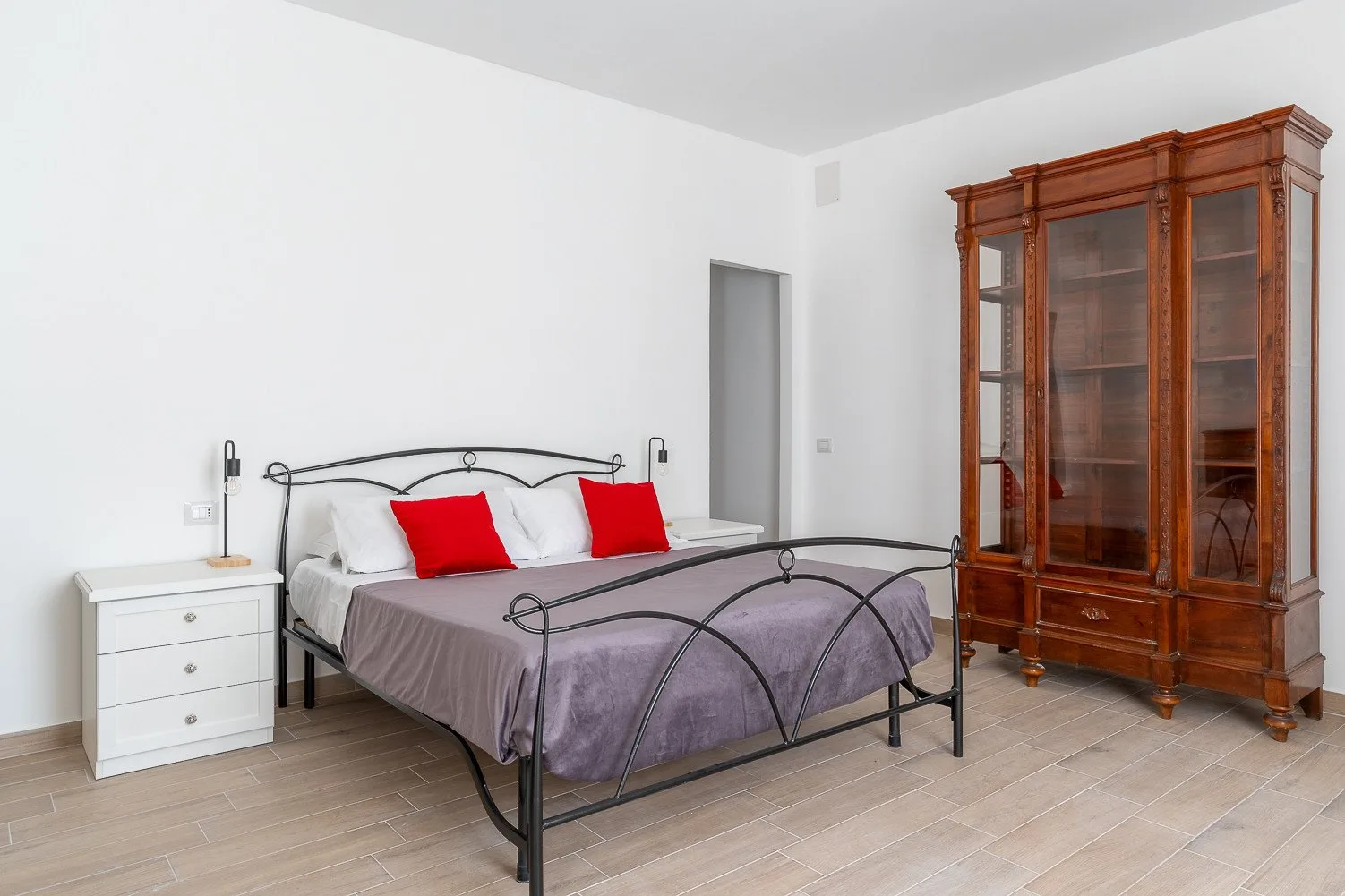 Un dormitorio con una cama de metal, mesitas blancas y una vitrina de madera.