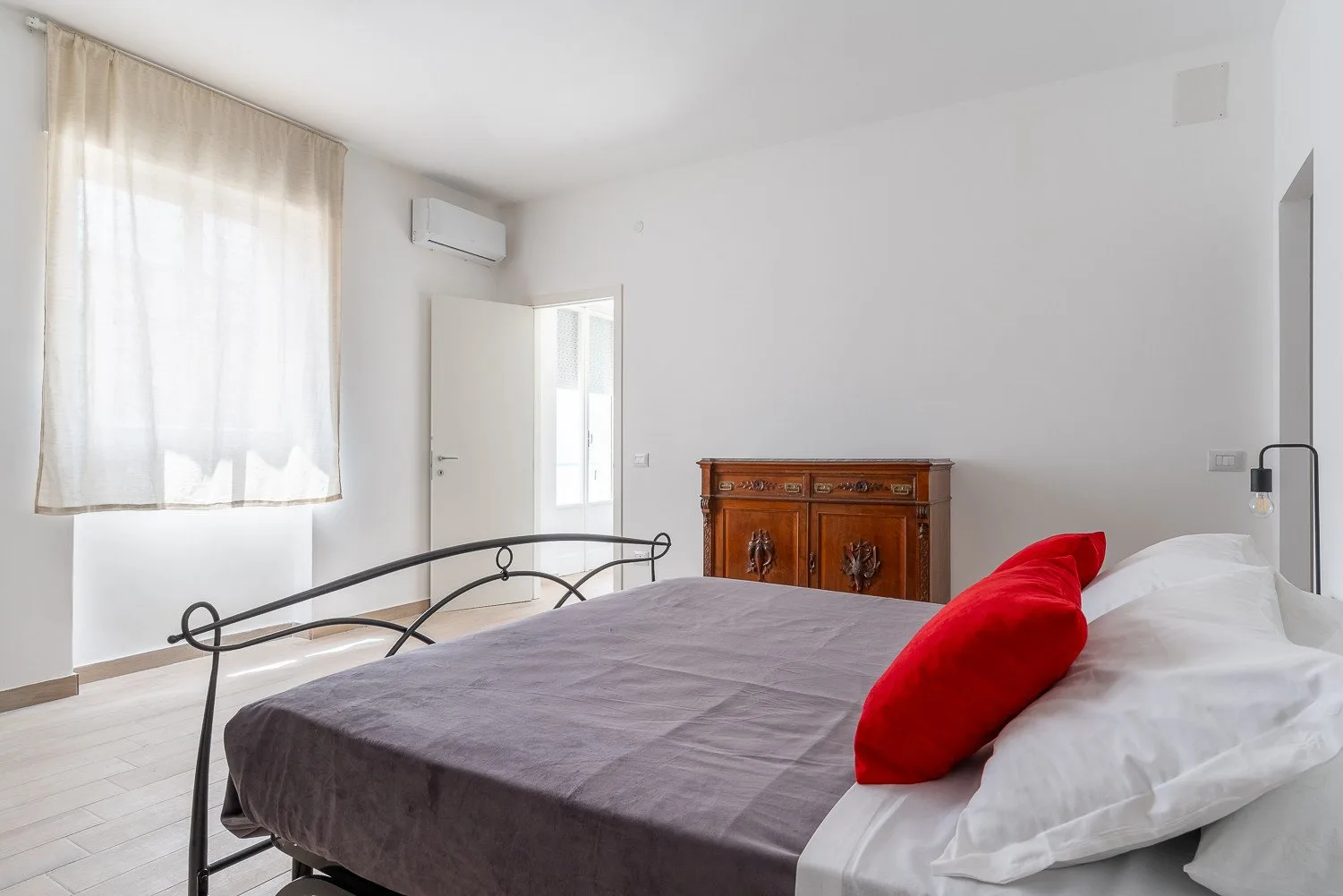 Un dormitorio con una cama vestida con ropa de cama gris y blanca y un cojín rojo.