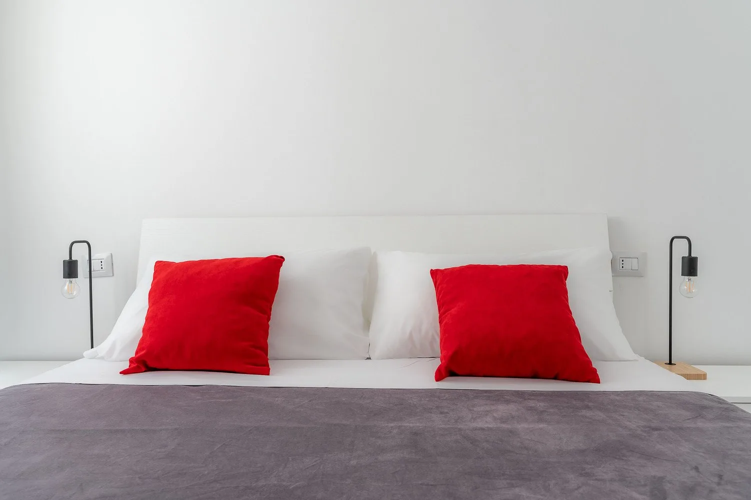 Una cama con almohadas blancas, cojines rojos y una colcha gris.