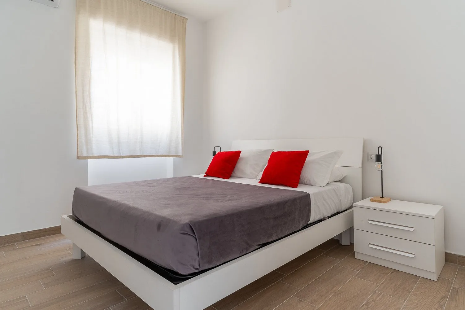 Un dormitorio luminoso con una cama vestida con ropa de cama gris y cojines rojos.