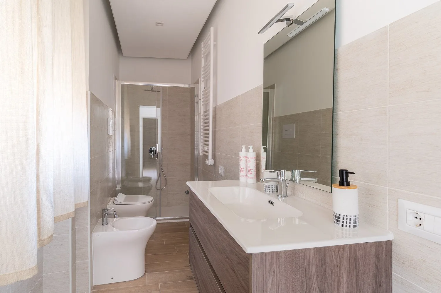 Un baño luminoso con un lavabo largo, un lavamanos y una ducha de vidrio.