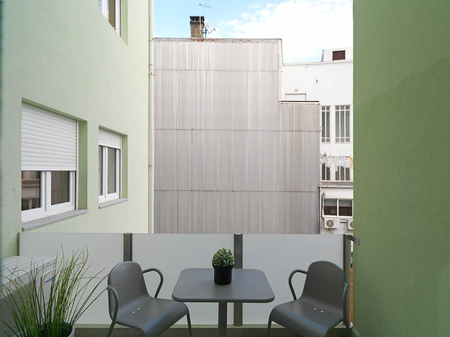 A small balcony with a table and seating faces neighboring apartment buildings.