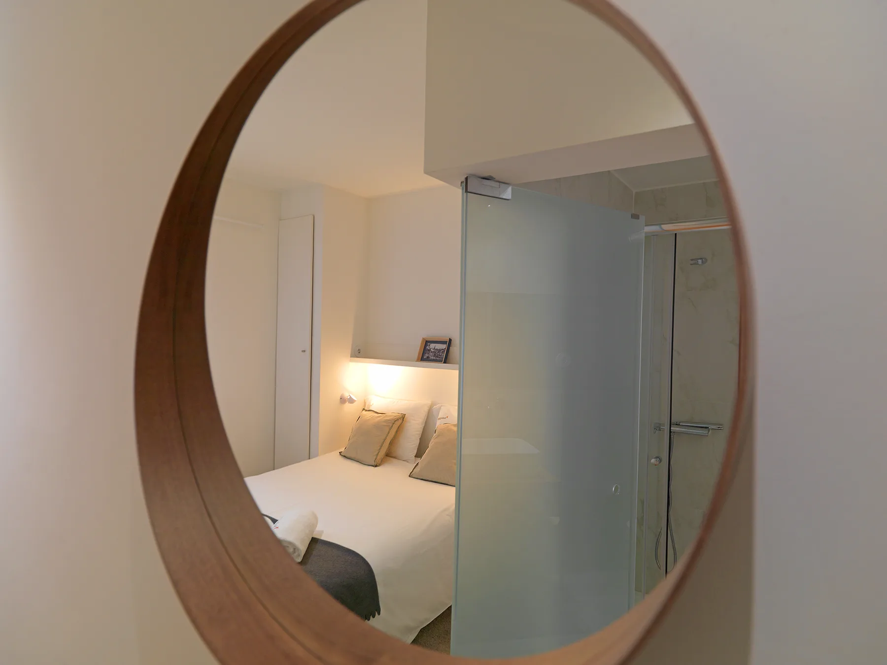 Dormitorio con una cama junto a una ducha de cristal, visto en un espejo redondo.