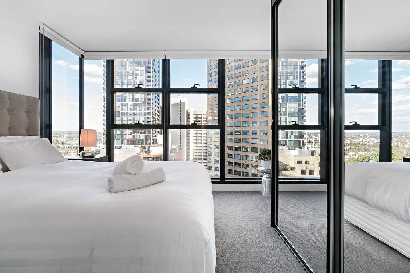 A bright bedroom with floor-to-ceiling windows overlooking nearby buildings.