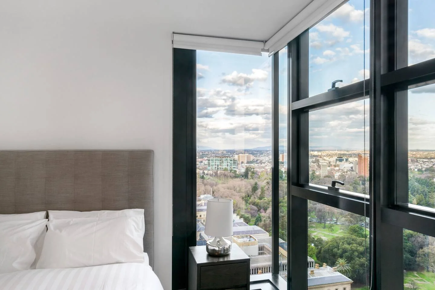 Dormitorio con una cama junto a grandes ventanas con vistas a edificios y vegetación.
