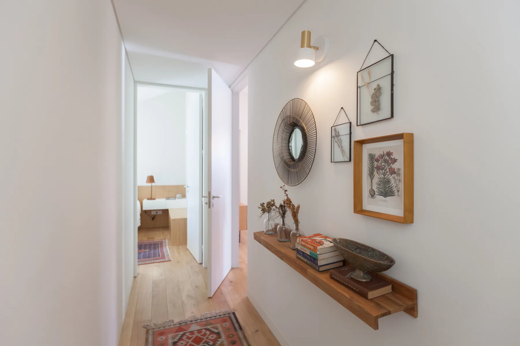 A bright apartment hallway features framed artwork, a round mirror, and a narrow wooden console.