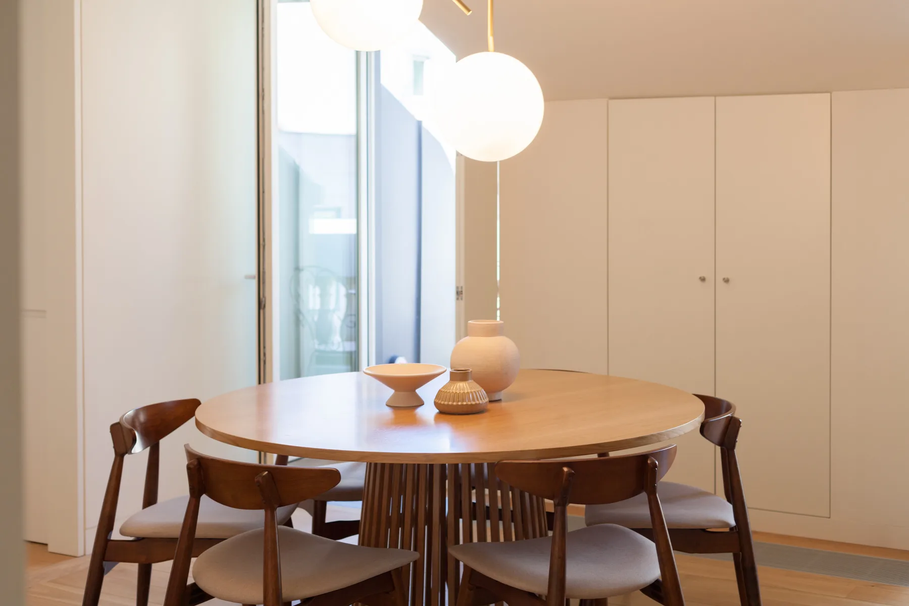 A dining area with a round wooden table and chairs beside tall windows.