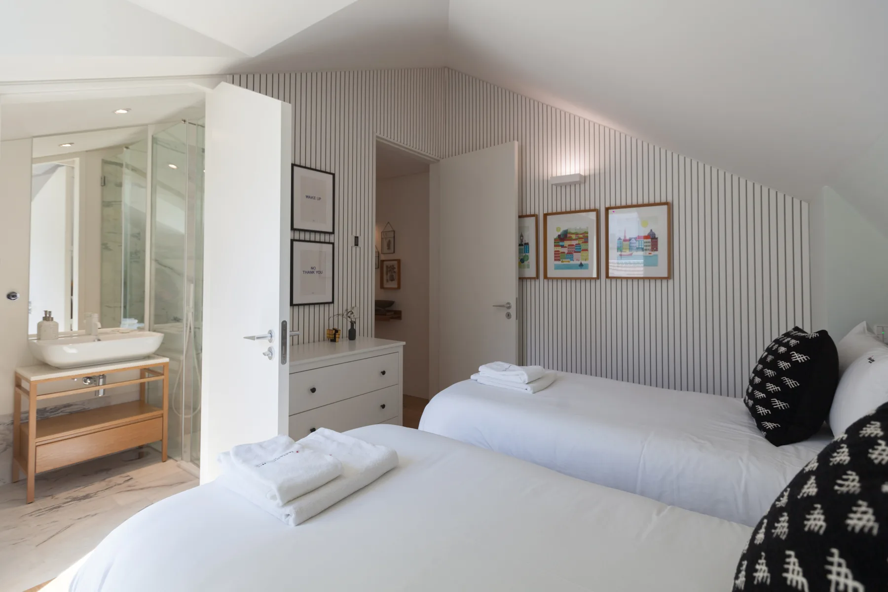 A bright bedroom with two beds, striped walls, and an open bathroom area.