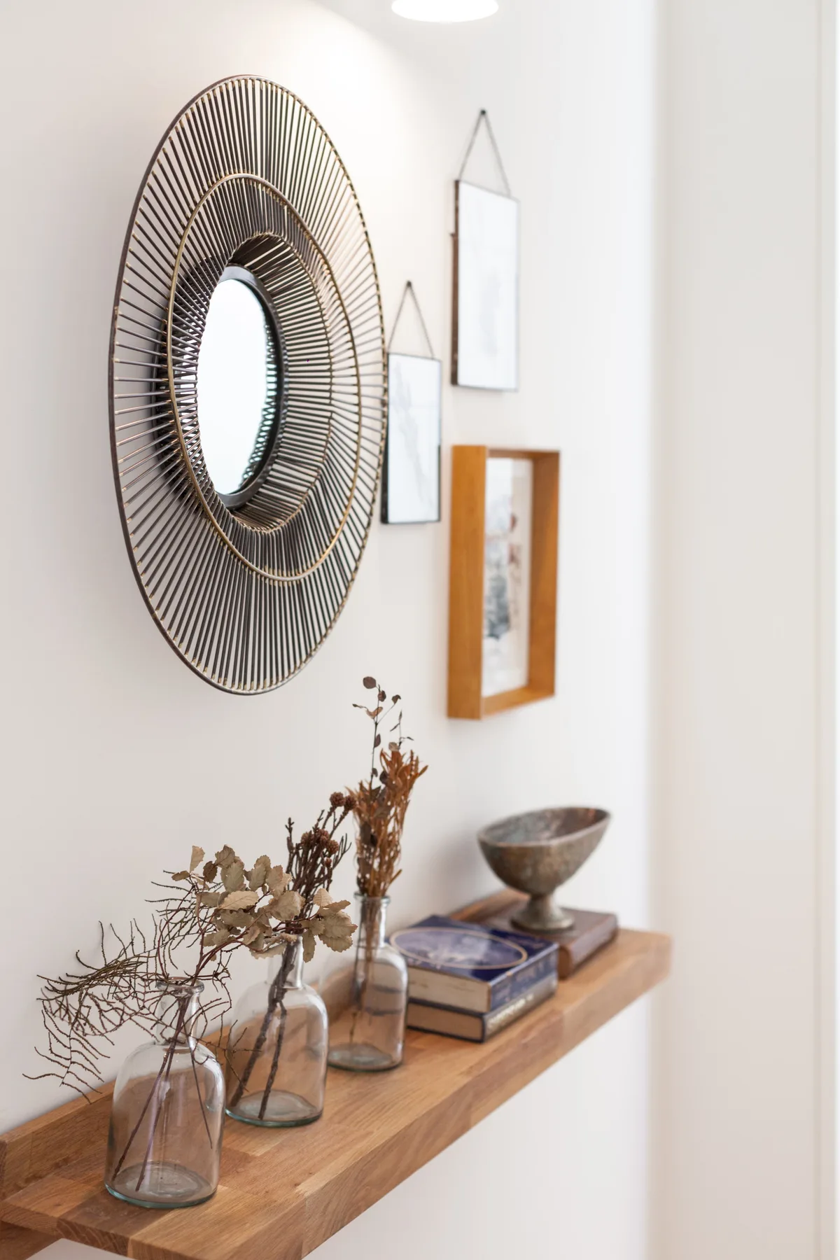 A bright interior wall has a round mirror, framed art, and a wooden display shelf.