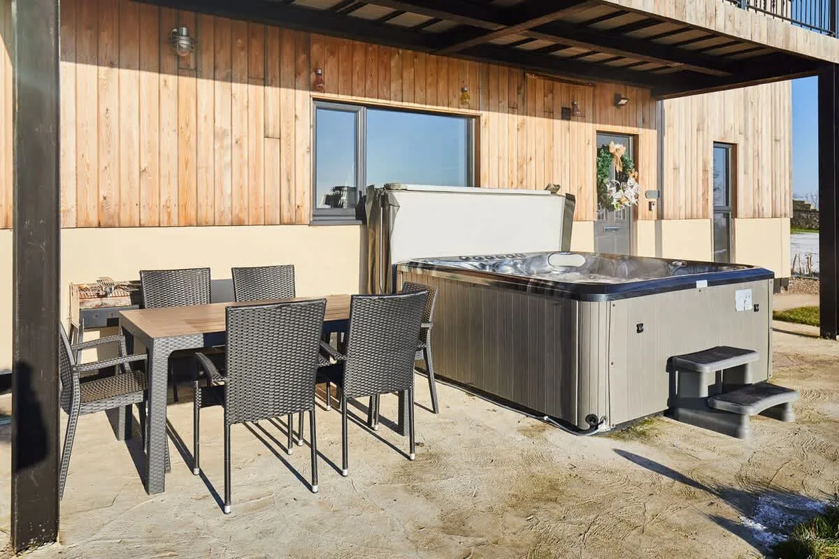An outdoor patio with a dining table and a hot tub beside a wooden-sided building.