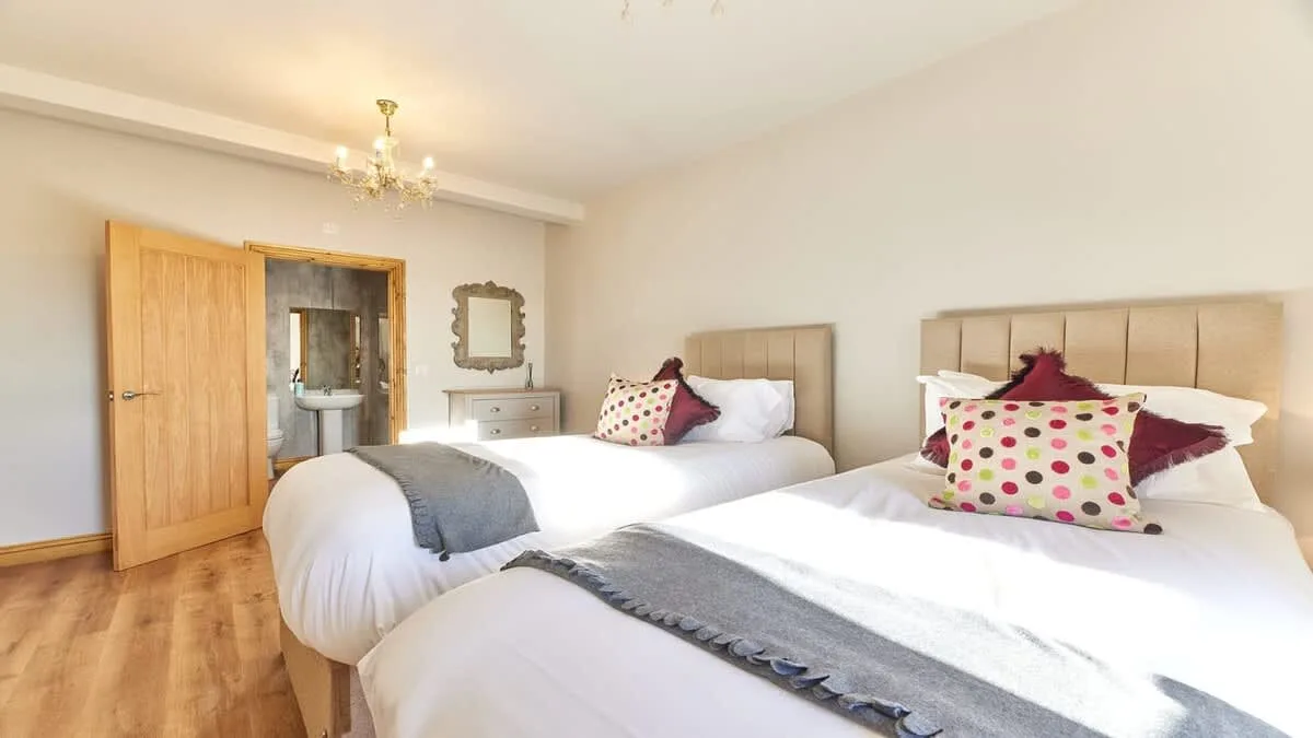 A bright bedroom with two beds dressed in white linens and colorful cushions.