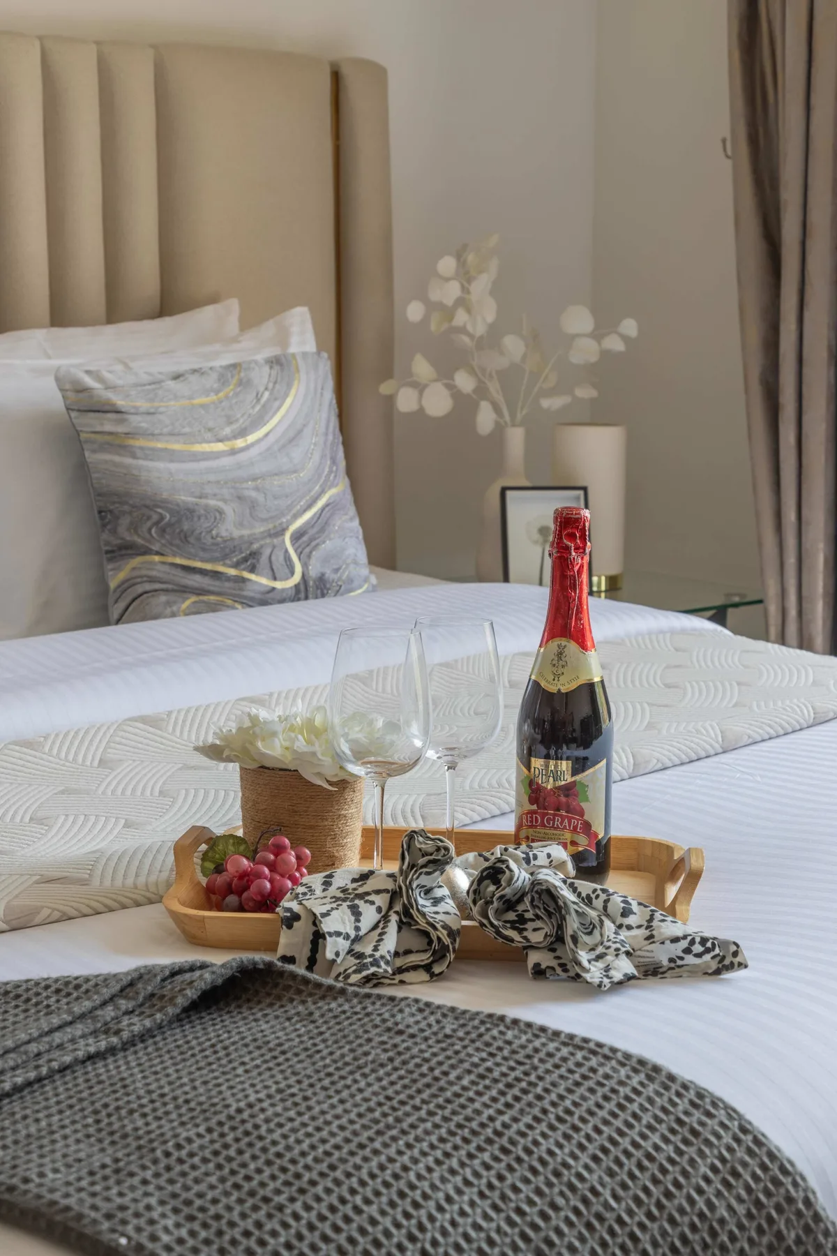 A neatly made bedroom bed is arranged with wine, glasses, fruit, and folded linens.