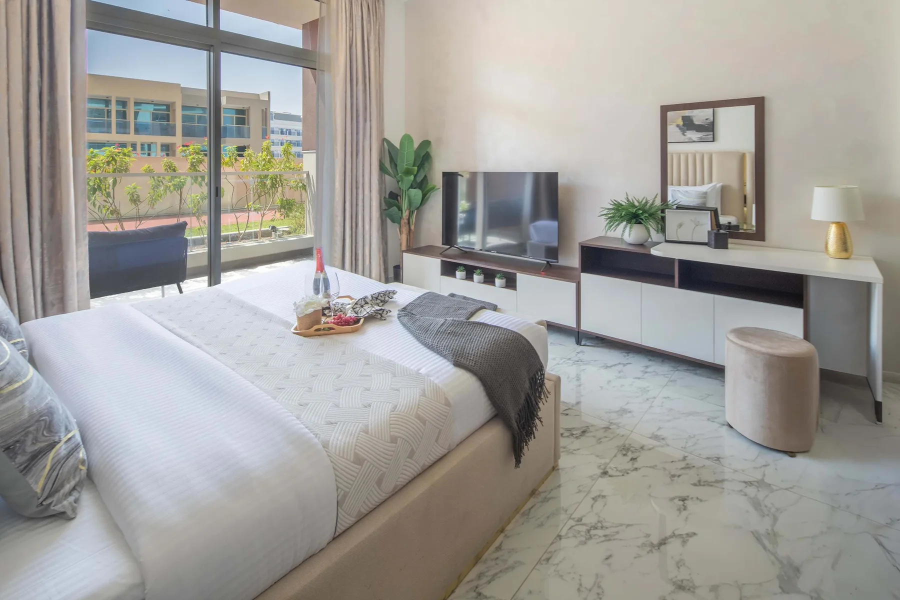 A bright bedroom with a bed, television, and large windows overlooking greenery.