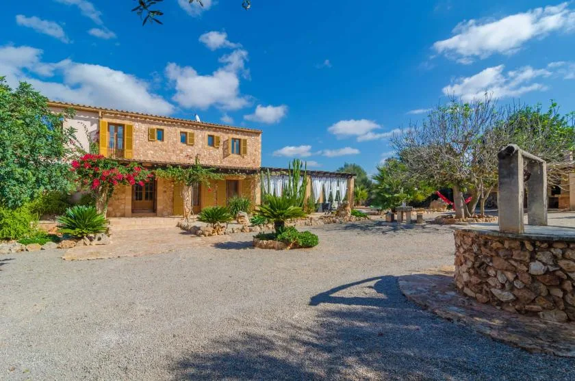 Una casa de piedra da a un amplio patio de grava con árboles y plantas con flores.