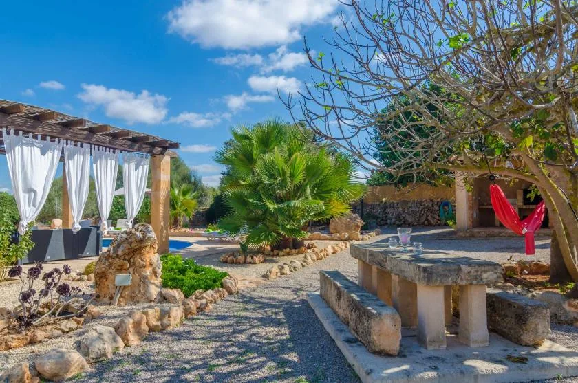 Un patio ajardinado al aire libre cuenta con asientos de piedra, plantas y una pérgola con sombra.