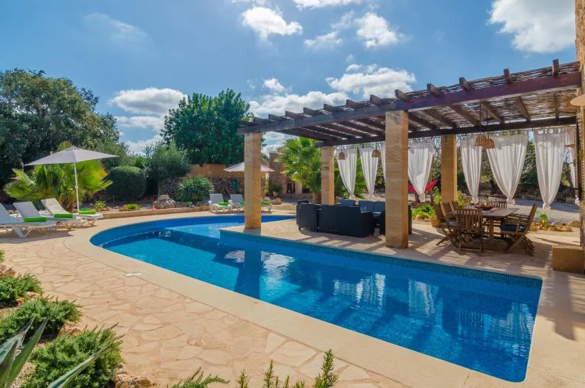 Una piscina al aire libre junto a una pérgola sombreada con asientos y cortinas.