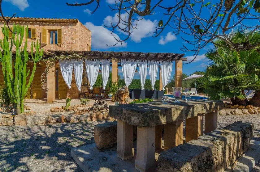 Un patio de piedra con una mesa larga al aire libre está junto a una pérgola con cortinas blancas.