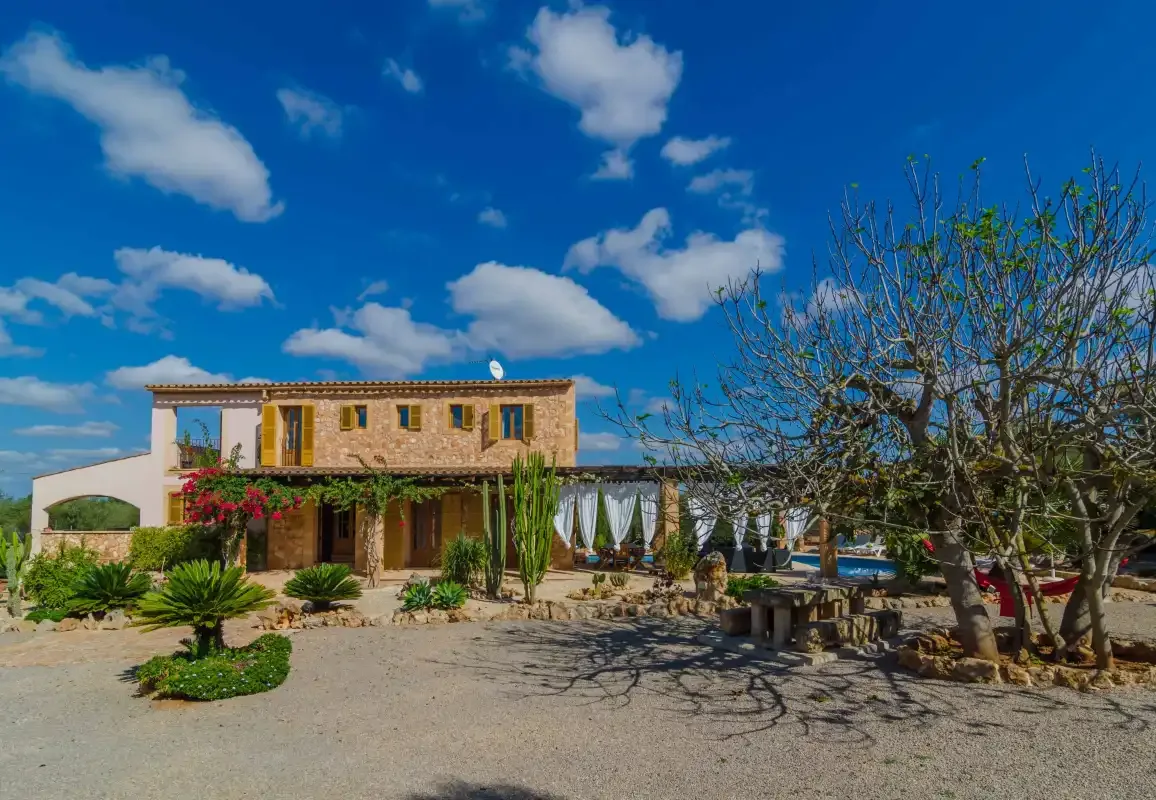 Una casa de dos plantas está entre árboles y arbustos bajo un cielo azul con nubes.