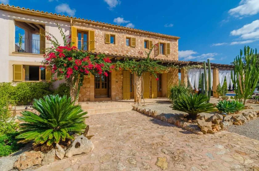 Una casa de piedra con patio pavimentado, enredaderas con flores y plantas desérticas.