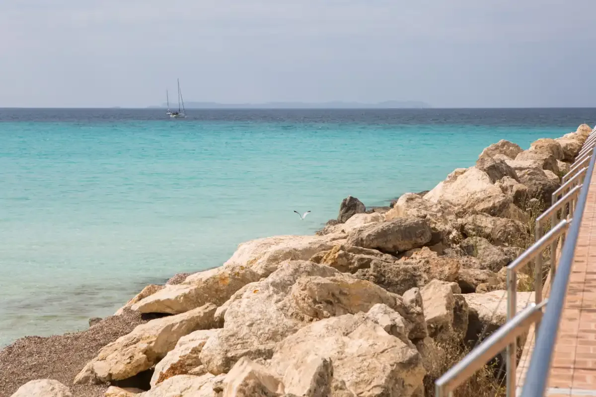 Una costa rocosa bordea aguas turquesas y claras, con un velero mar adentro.