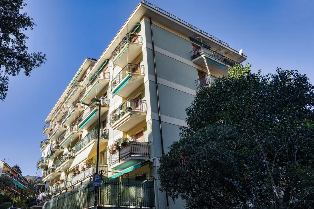 Un edificio de apartamentos de varios pisos con balcones se alza junto a árboles frondosos bajo un cielo despejado.