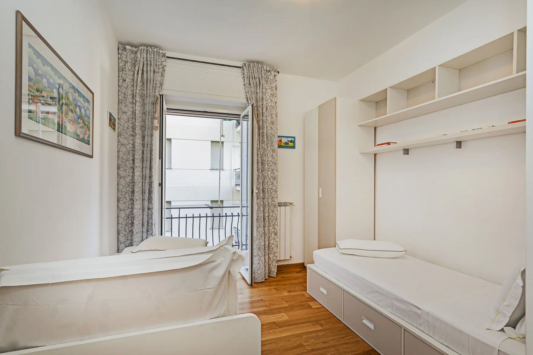 Un dormitorio luminoso con dos camas, estantes abiertos y una puerta al balcón.