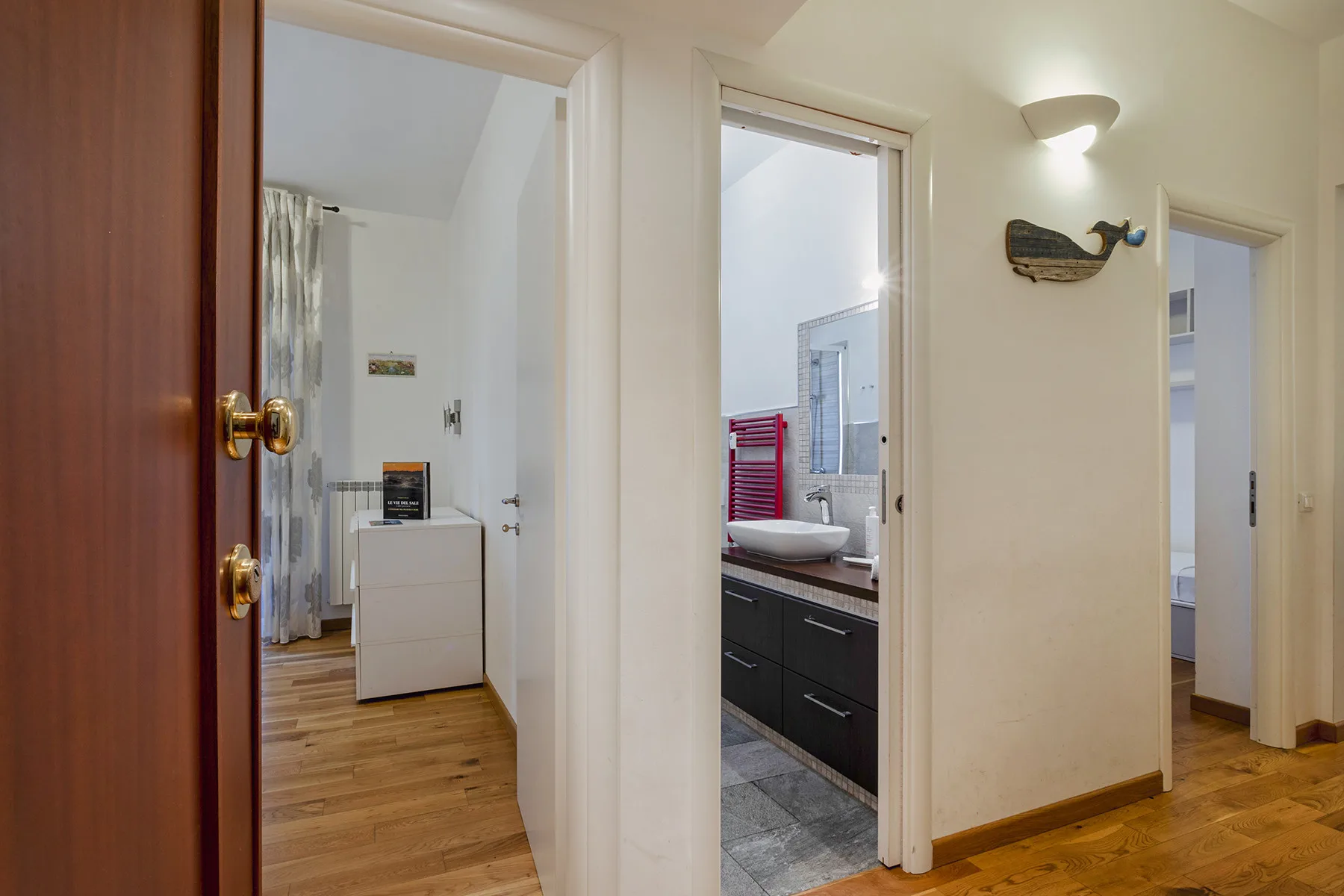 Un pasillo luminoso conecta varias puertas abiertas, con suelo de madera y una lámpara de pared.