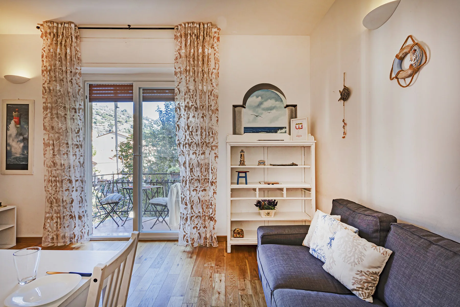 Una sala de estar con sofá gris, estantería y puertas de vidrio hacia un balcón.