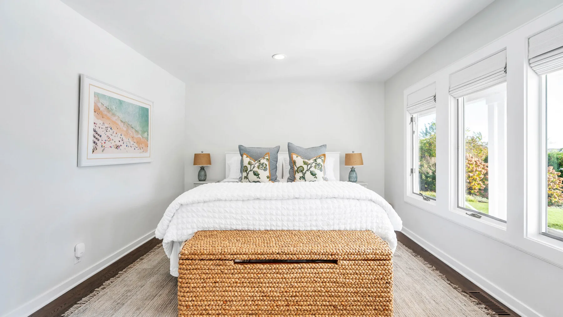 Un dormitorio luminoso con una cama, un banco tejido y ventanas grandes.