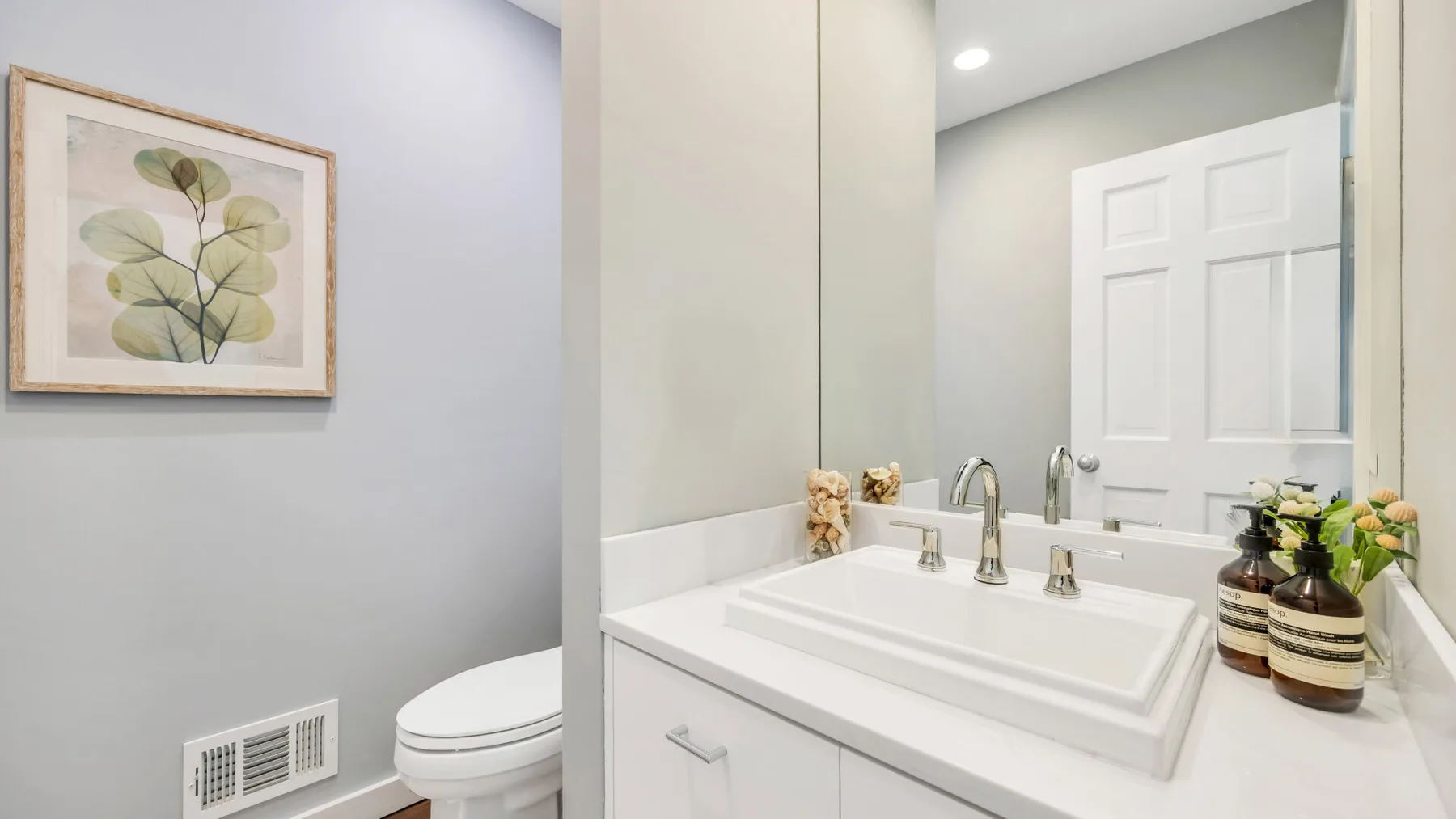 Un tocador de baño con lavabo blanco, espejo y lámina botánica enmarcada.