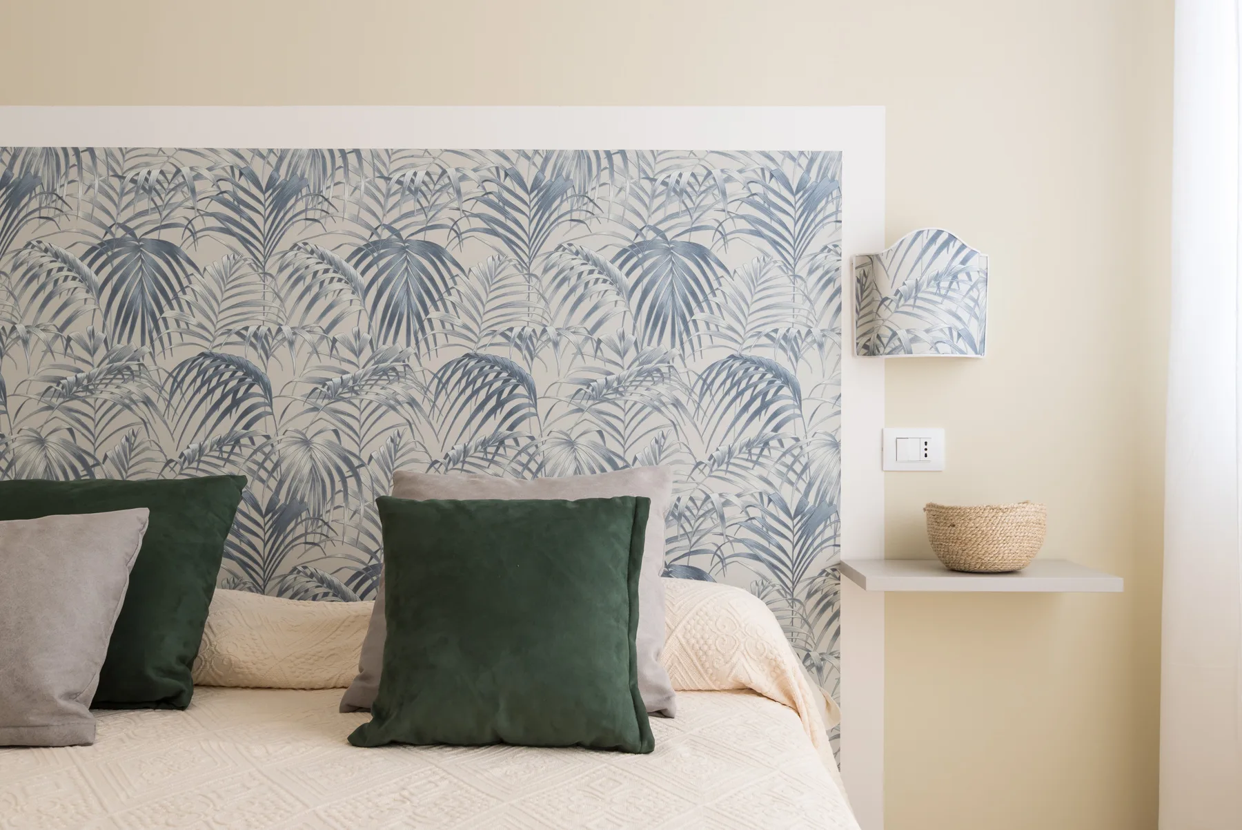 A bedroom with a bed against a palm-patterned wall and green pillows.