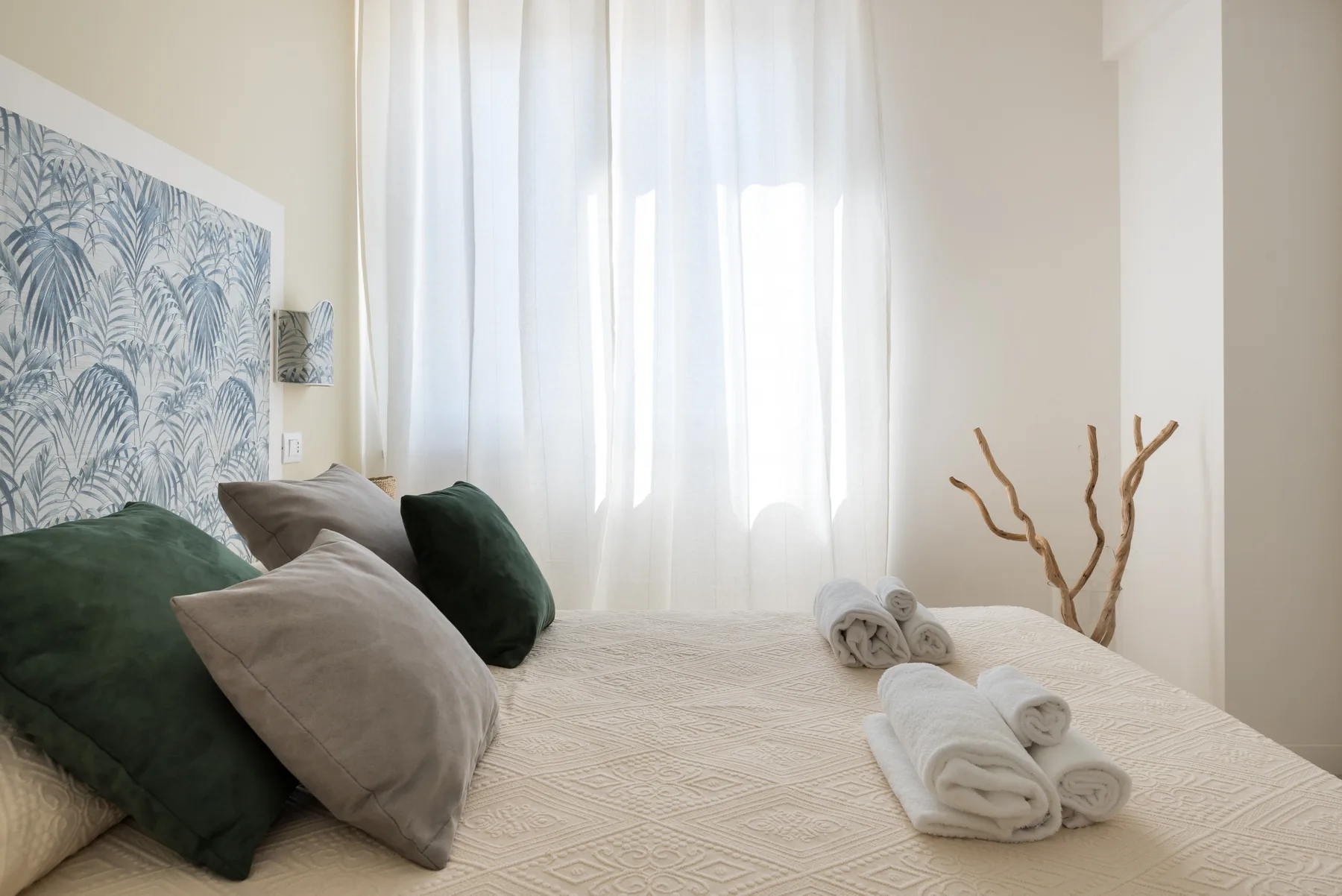 A bright bedroom with a neatly made bed, folded towels, and sheer curtains.