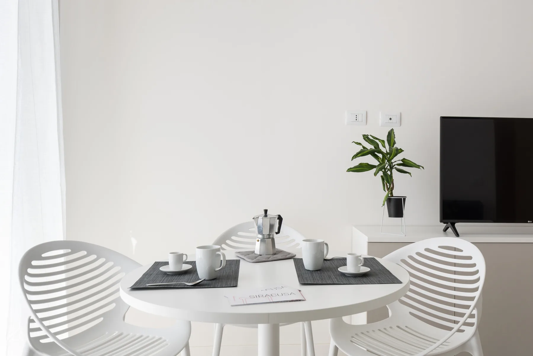 A bright dining area has a round table set with cups beside a television and potted plant.