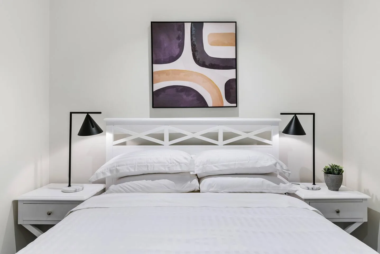 Un dormitorio luminoso con una cama blanca, mesitas de noche a juego y arte abstracto enmarcado.