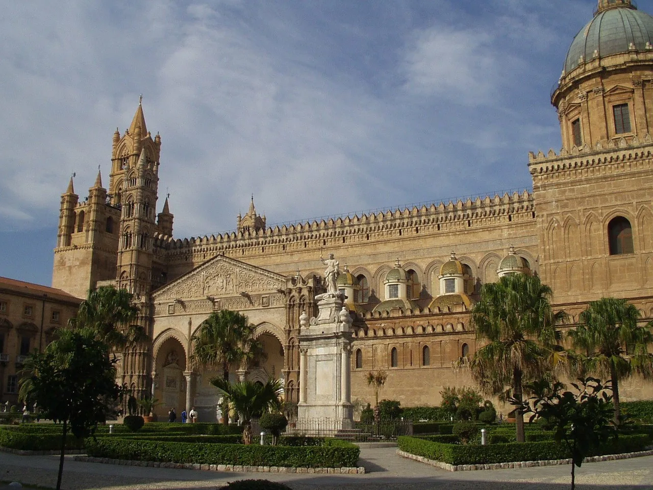 A grand historic stone cathedral façade rises above a landscaped plaza with palm trees.