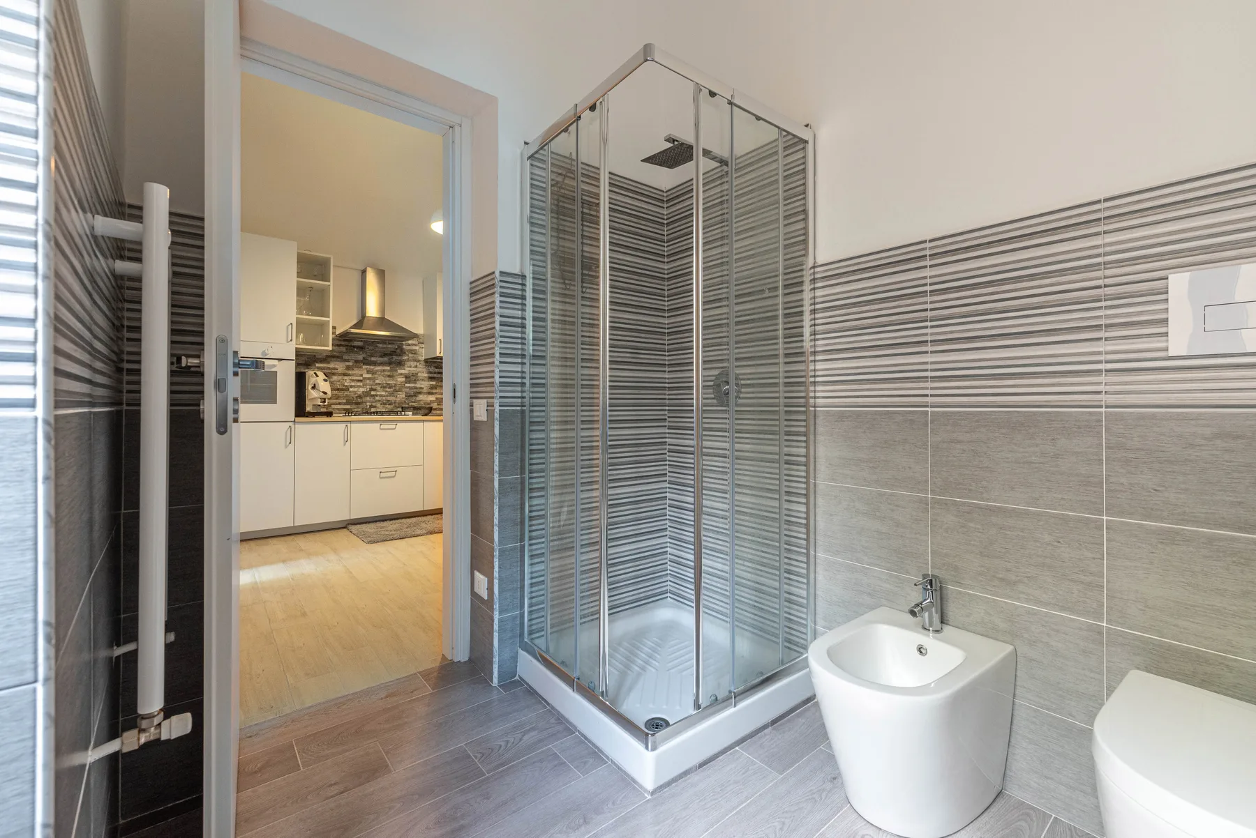 A tiled bathroom with a glass shower and a doorway to the kitchen.