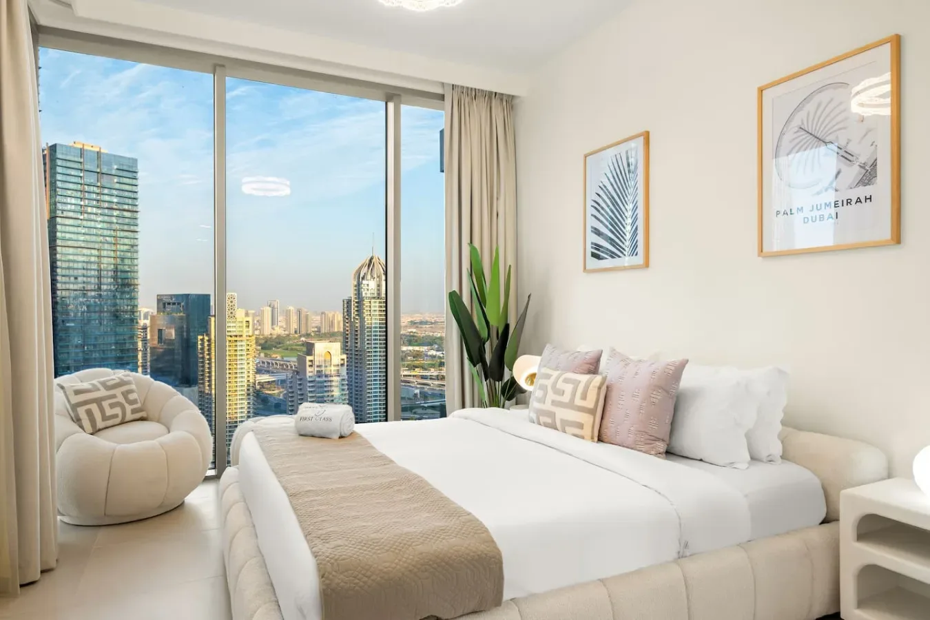 A bright bedroom with a large bed, seating, and floor-to-ceiling windows overlooking the city.