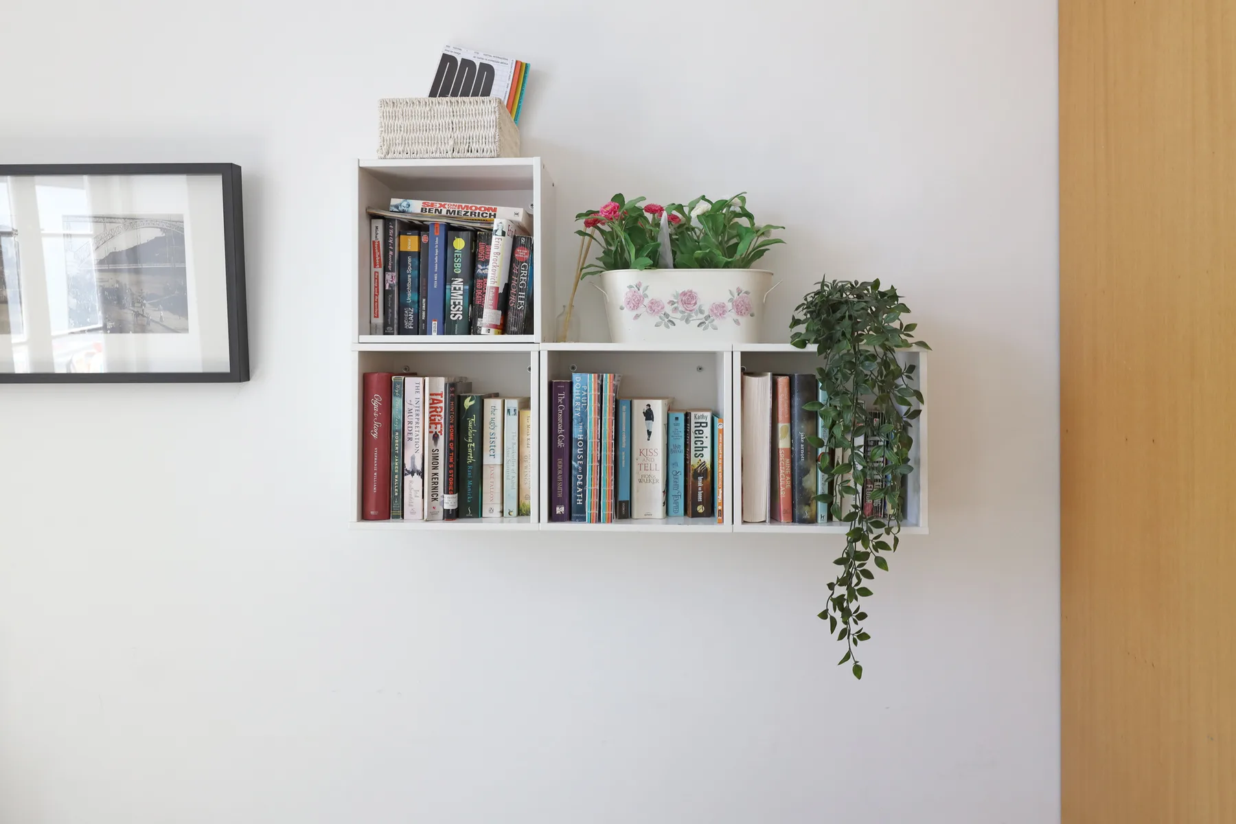 A wall-mounted bookshelf holds books and potted plants beside a framed picture.
