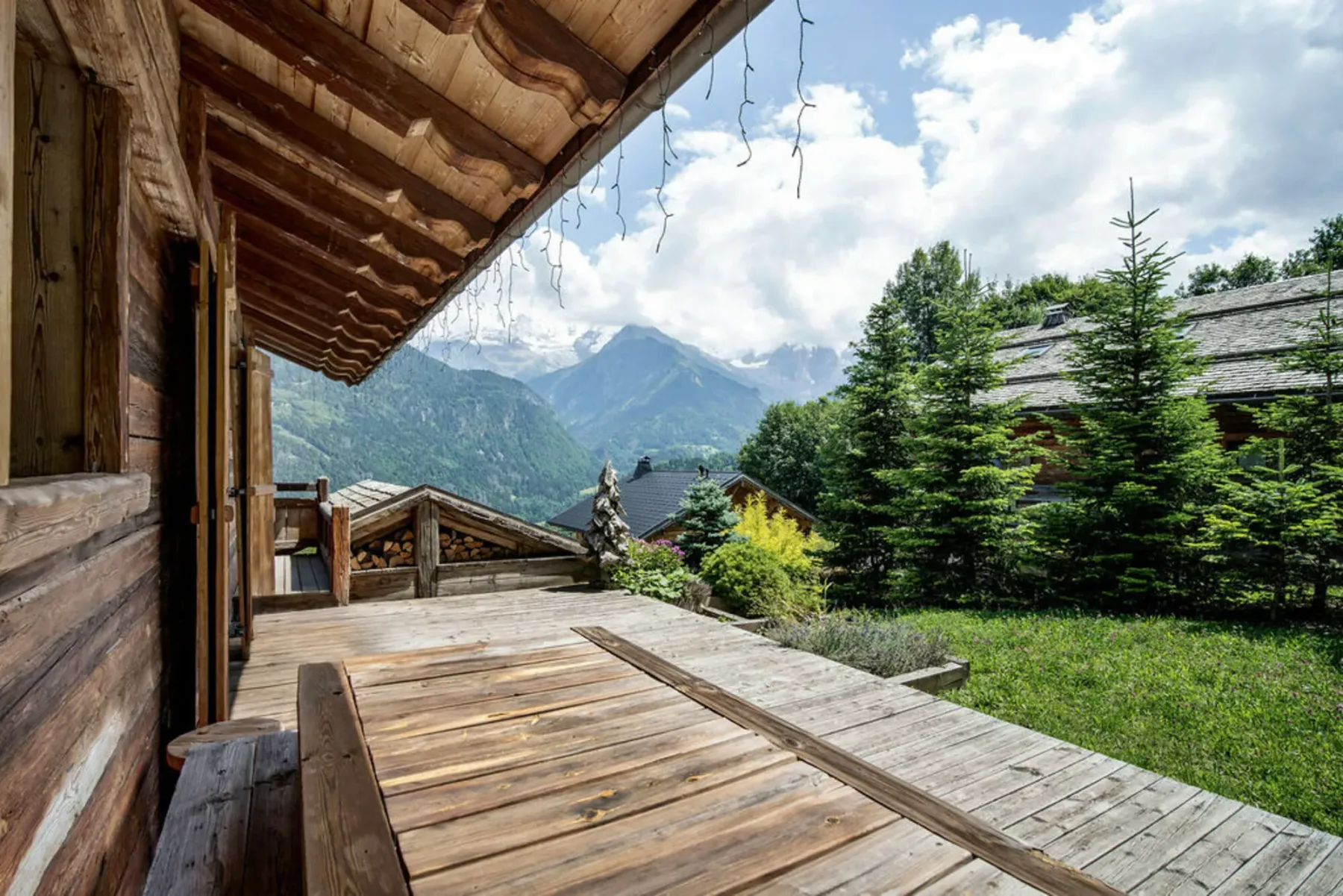 A wooden terrace overlooks green trees and a mountain valley beneath a cloudy sky.