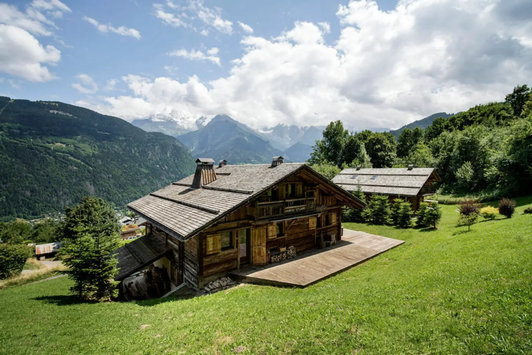 A wooden cabin sits on a grassy hillside with mountains in the background.