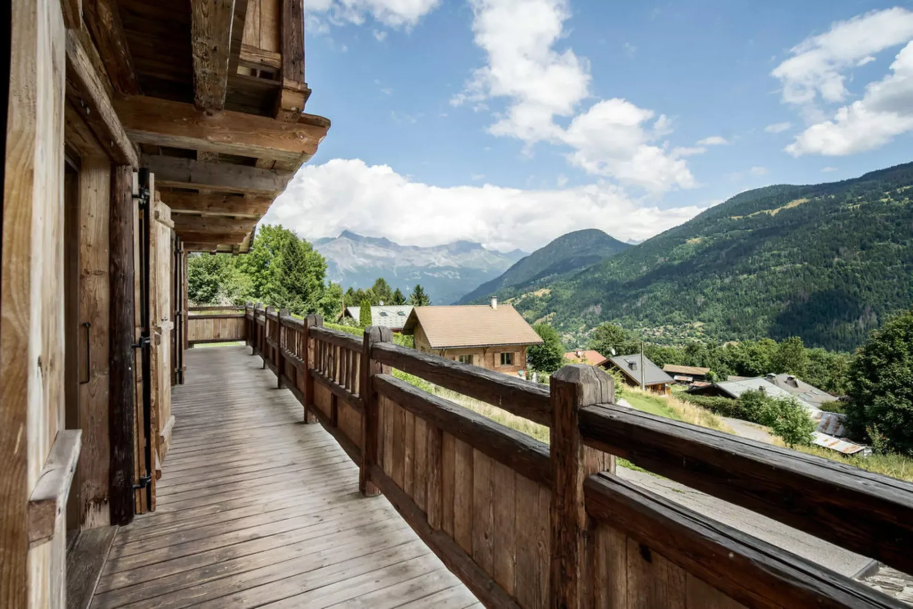 A wooden balcony overlooks mountain slopes and a valley beneath a partly cloudy sky.