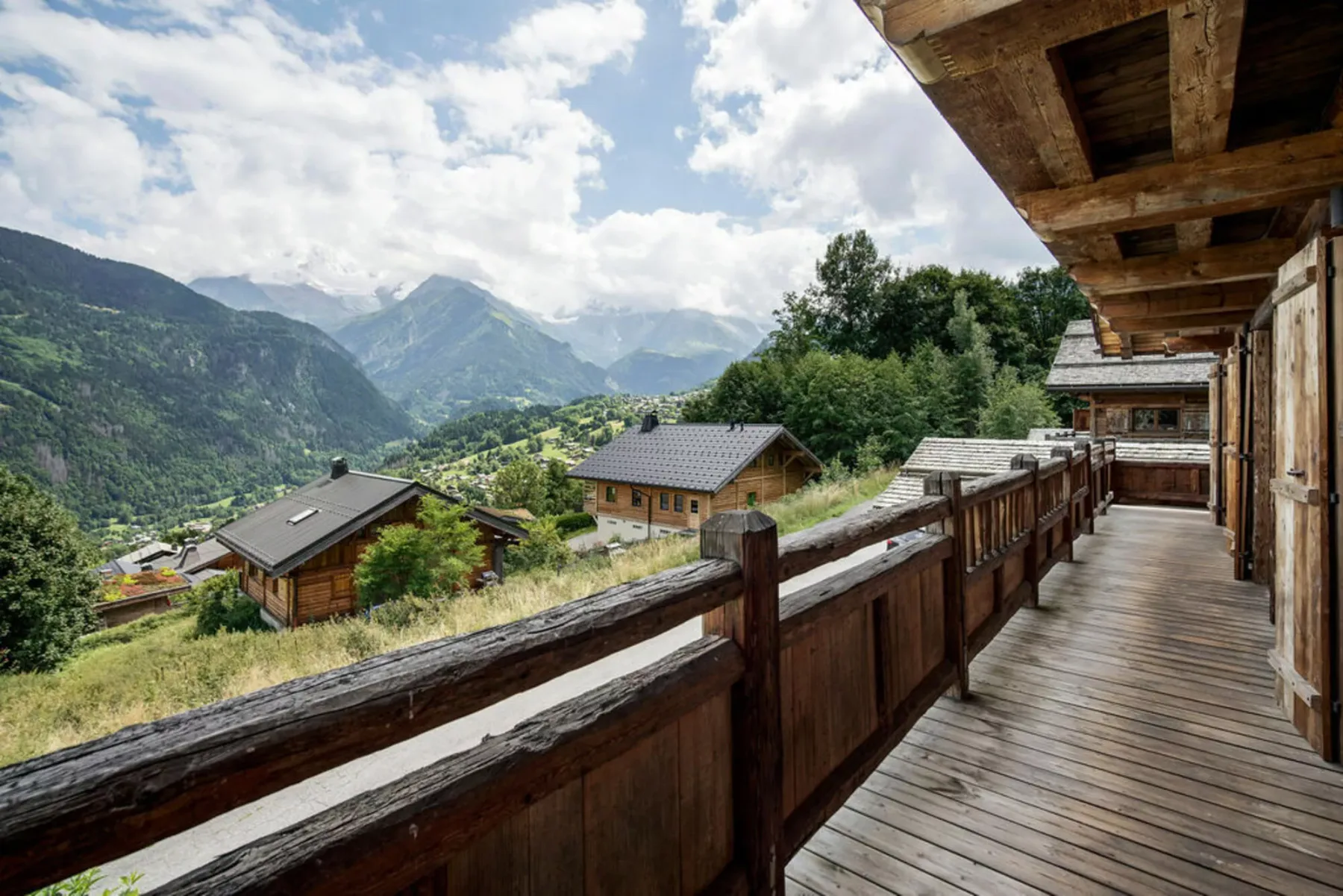 A wooden balcony overlooks mountain slopes and scattered houses beneath a cloudy sky.