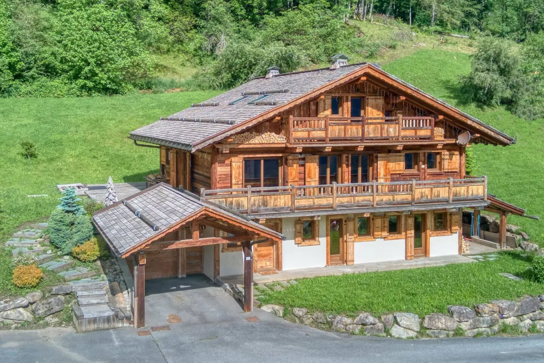 A large wooden chalet stands on a grassy hillside, with a covered driveway beside it.