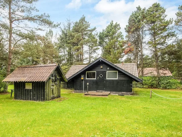 A dark cottage stands on a grassy lawn among tall trees.