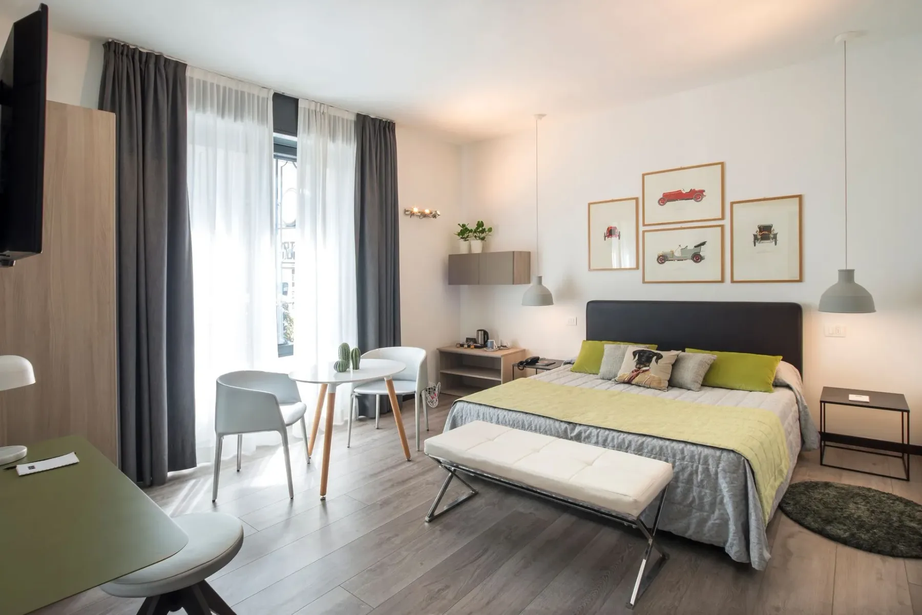 Bright hotel room with a bed, table, chairs, artwork, and wood flooring.