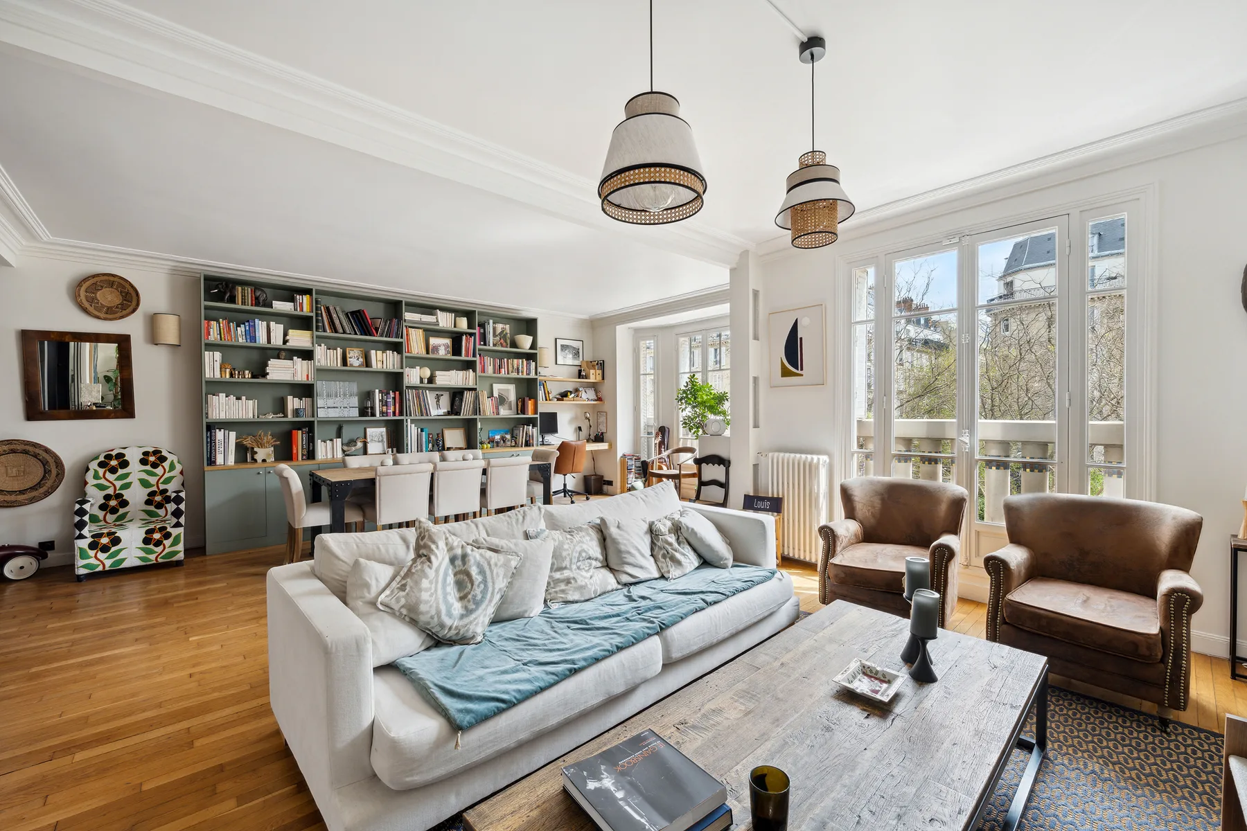 Una sala de estar luminosa con sofá, sillones y estanterías junto a grandes ventanas.