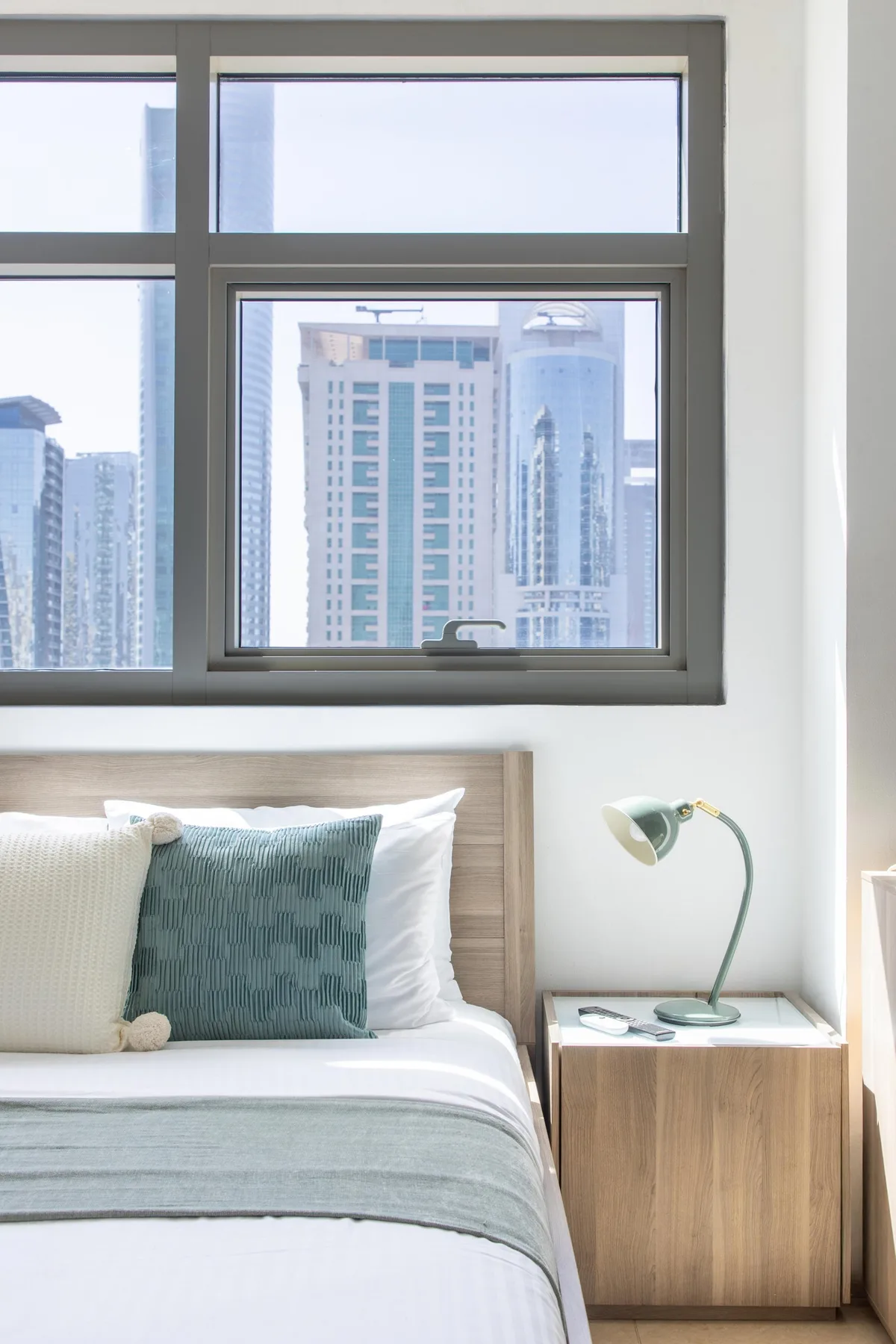 A bedroom with a neatly made bed beside a window overlooking city buildings.