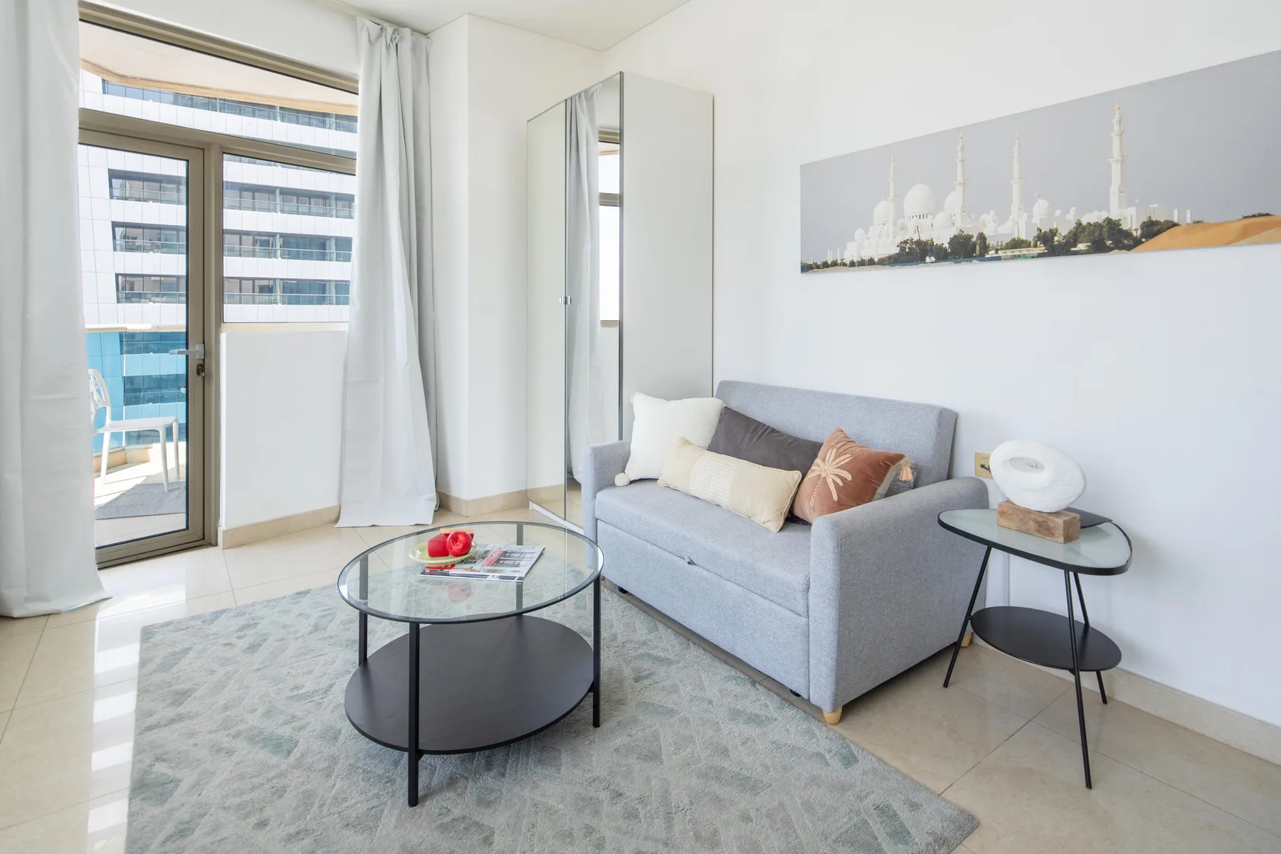 A bright living room has a gray sofa, glass coffee table, and balcony doors.